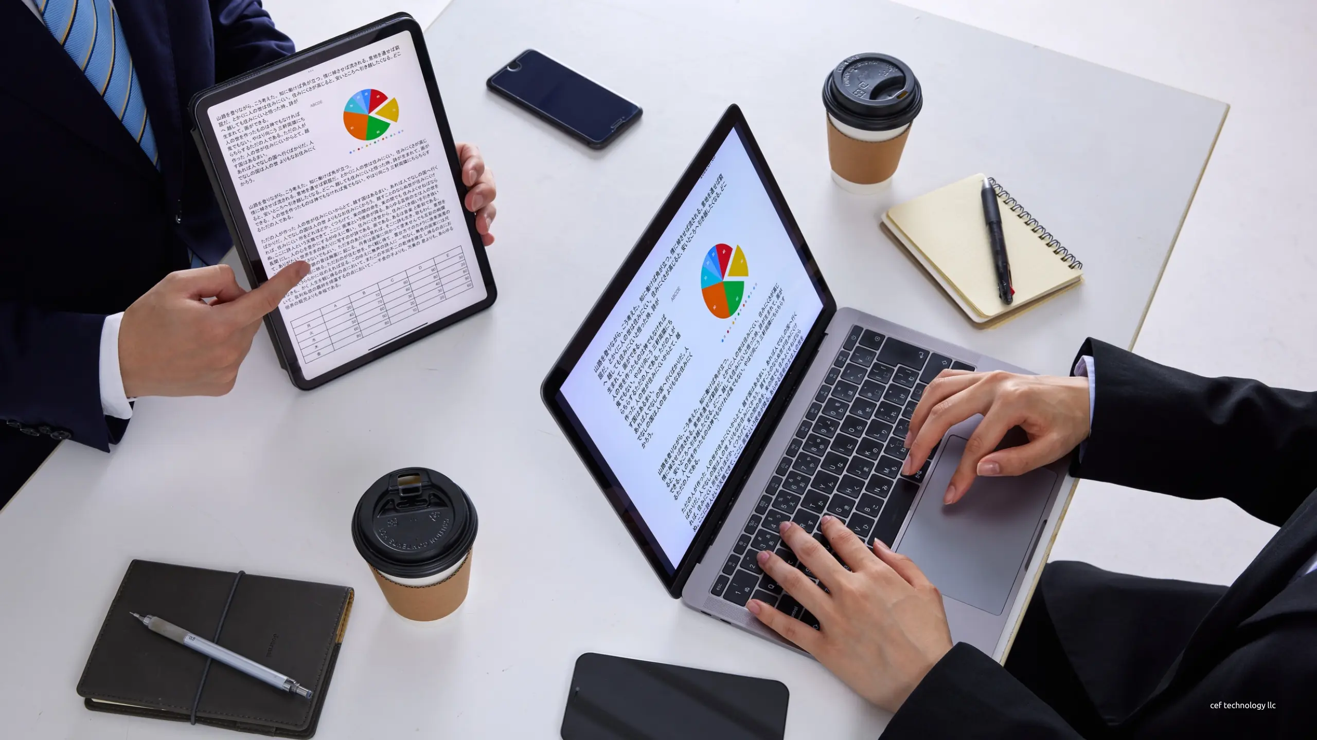 Two people at a white table working through charts on a tablet and a laptop, seen from above.