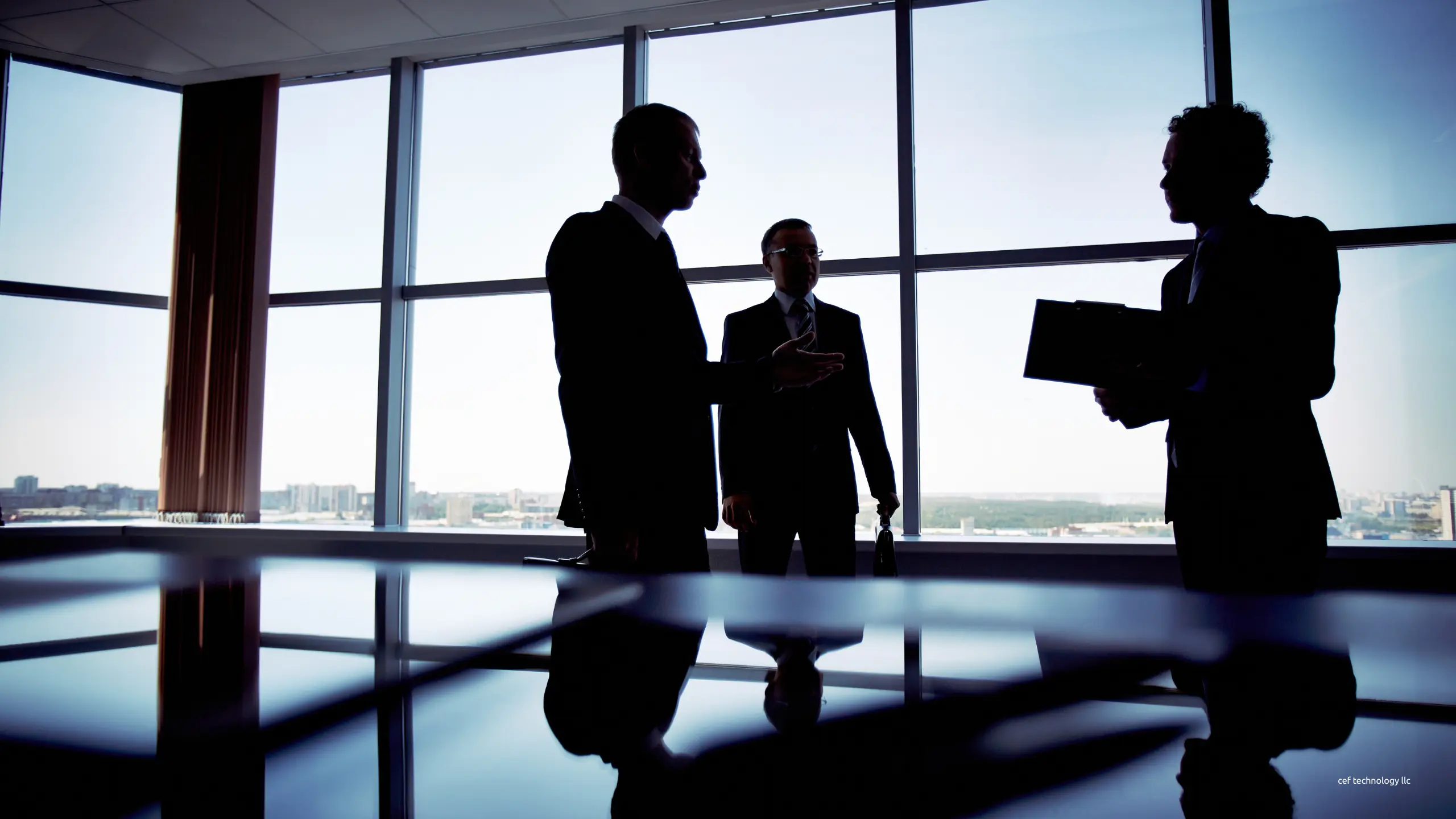 Three people in suits talking by a floor to ceiling office window, seen in silhouette against the city beyond.
