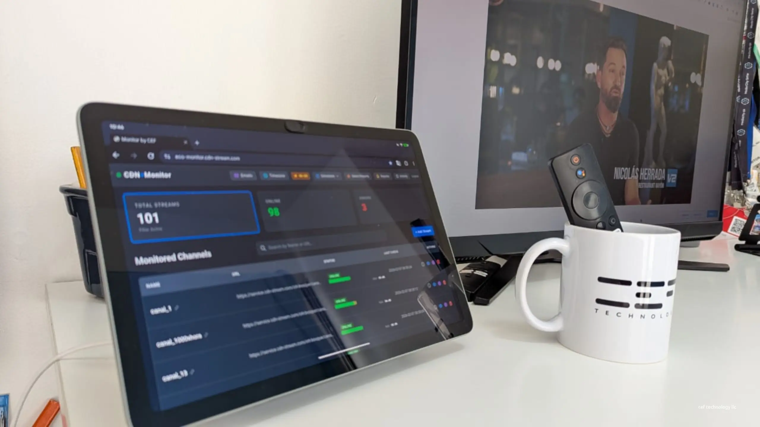 A tablet on a desk showing CEF’s channel monitoring panel, with a television playing behind it and a CEF mug beside it.
